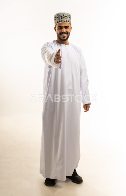 إلقاء التحيه والسلام, بورتريه لشاب عربي خليجي عماني مبتسم يرتدي الدشداشة و الكمه يقوم بمد كفه للمصافحة ينظر الى الكاميرا بإيماءات السرور، التعارف و الصداقه وتكوين علاقات, مفهوم الاستقبال والترحيب، صورة الجسم بالطول الكامل، خلفية بيضاء