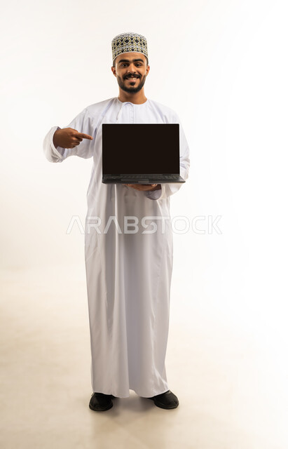 Using modern technologies and devices, the concept of remote business management, a portrait of a smiling young Gulf Arab Omani man wearing casual clothes holding a laptop in his hand, looking at the camera with gestures of pleasure, pointing his index finger at a blank screen, a full-length body image, white background