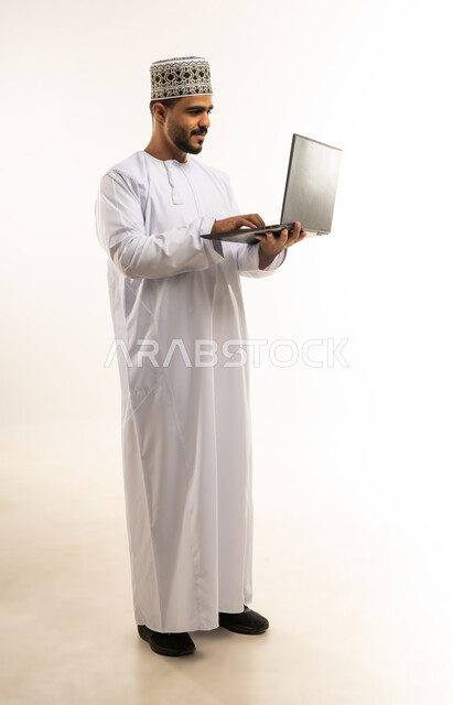 اعداد التقارير الخاصة بالعمل، مفهوم إدارة الأعمال عن بعد، بورتريه لشاب عربي خليجي عماني مبتسم يرتدي الدشداشة و الكمه يحمل بيده جهاز اللابتوب المحمول، استخدام التقنيات والأجهزة الحديثة لإنجاز المهام، إجراء مكالمات فيديو, صورة الجسم بالطول الكامل، خلفية بيضاء