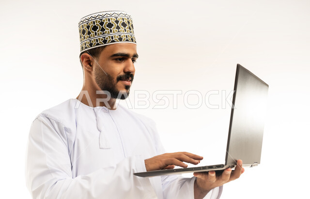 مفهوم إدارة الأعمال عن بعد، اعداد التقارير الخاصة بالعمل، بورتريه مقرب لشاب عربي خليجي عماني مبتسم يرتدي الدشداشة و الكمه يحمل بيده جهاز اللابتوب المحمول، استخدام التقنيات والأجهزة الحديثة لإنجاز المهام، إجراء مكالمات فيديو, خلفية بيضاء