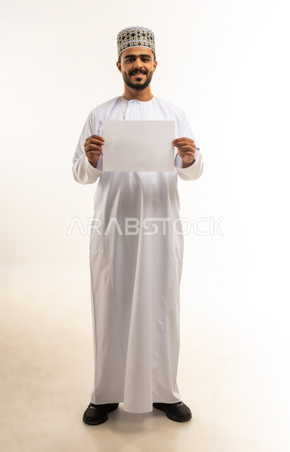 موك اب مستطيل أبيض، استخدام لوحات توضيحية تعريفية، بورتريه لشاب عربي خليجي عماني مبتسم يرتدي الدشداشة والكمه يحمل بيده لوحه بيضاء مستطيلة فارغة ينظر الى الكاميرا بإيماءات الفرح السرور، إعلانات وعروضات تسويقية، صورة الجسم بالطول الكامل، خلفية بيضاء