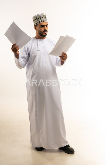 اعداد التقارير الخاصة بالعمل، بورتريه لشاب عربي خليجي عماني مبتسم يرتدي الدشداشة و الكمه يحمل بيده مجموعة من الاوراق البيضاء، مراجعة ومتابعة الخطط والمشاريع المكتبية بإيماءات التركيز، التدقيق في قراءة الملاحظات، صورة الجسم بالطول الكامل، خلفية بيضاء