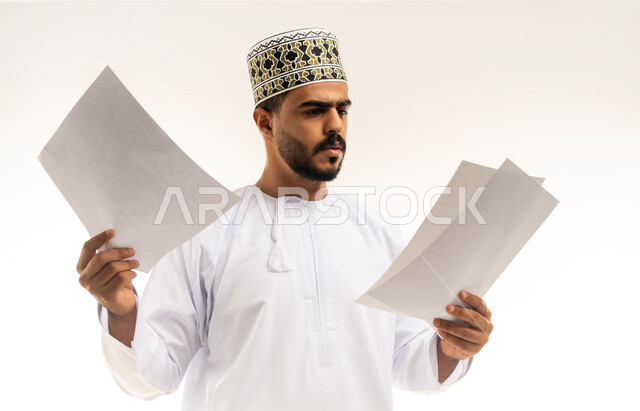 التدقيق في قراءة الملاحظات، اعداد التقارير الخاصة بالعمل، بورتريه لشاب عربي خليجي عماني مبتسم يرتدي الدشداشة و الكمه يحمل بيده مجموعة من الاوراق البيضاء، مراجعة ومتابعة الخطط والمشاريع المكتبية بإيماءات التركيز، خلفية بيضاء