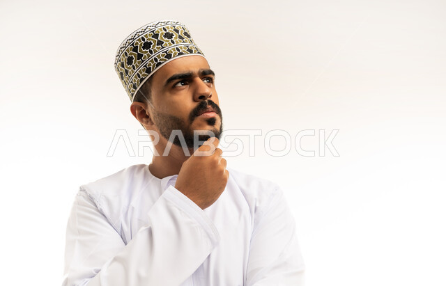 التأمل والتخطيط والتمعن بأمر محدد, بورتريه مقرب لرجل عربي خليجي عماني يرتدي الدشداشة و الكمه يضع يديه على ذقنه بإيماءات التفكير, ايجاد الافكار والحلول, خلفية بيضاء