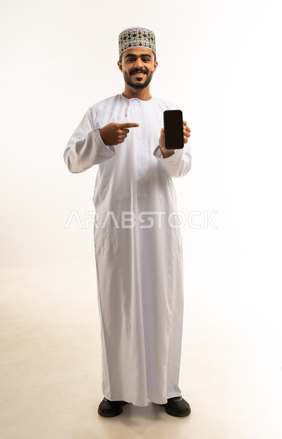 استخدام الوسائل التقنية الحديثة، عرض شاشة سوداء فارغة على الهاتف المحمول، النظر إلى الكاميرا بايماءات الفرح والسرور، بورتريه لشاب عربي خليجي عماني يرتدي الدشداشة والكمه يحمل الجوال ويشير اليه بإصبع السبابة، صورة لكامل الجسم، خلفية بيضاء