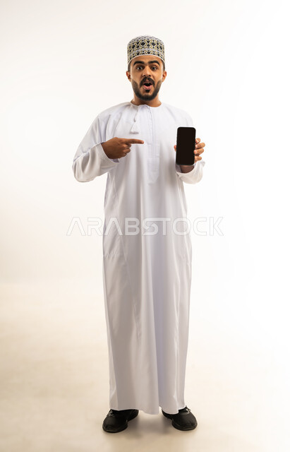 عرض شاشة سوداء فارغة على الهاتف المحمول، استخدام الوسائل التقنية الحديثة، النظر إلى الكاميرا بايماءات المفاجئة والتعجب، بورتريه لشاب عربي خليجي عماني يرتدي الدشداشة والكمه يحمل الجوال ويشير اليه بإصبع السبابة، صورة لكامل الجسم، خلفية بيضاء
