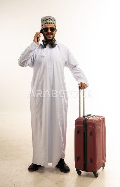 إجراء مكالمات هاتفية، الاستعداد لقضاء رحلة صيفية ممتعة, بورتريه لشاب عربي خليجي عماني مبتسم يرتدي الدشداشة والكمه والنظارة الشمسية يمسك حقيبة السفر والهاتف المحمول ينظر الى الكاميرا بإيماءات الفرح, صورة الجسم بالطول الكامل، خلفية بيضاء