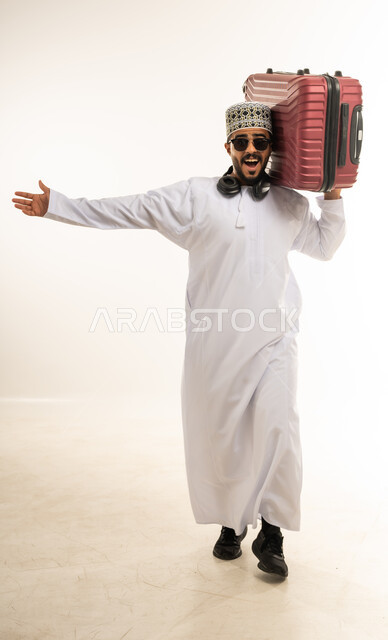 الذهاب في جولات ترفيهية في إجازة الصيف, تعابير الفرح والسرور، بورتريه لشاب عربي خليجي عماني مبتسم يرتدي الدشداشة والكمه والنظارة الشمسية يحمل حقيبة السفر على كتفه, الاستعداد لقضاء رحلة صيفية ممتعة, الشعور بالمتعة والحماس، صورة الجسم بالطول الكامل، خلفية بيضاء