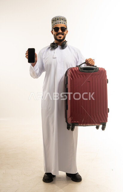 حجز اونلاين عبر تطبيقات وبرامج السياحة, بورتريه لشاب عربي خليجي عماني مبتسم يرتدي الدشداشة والكمة والنظارة الشمسية يحمل حقيبة السفر يمسك بيده الهاتف المحمول, الاستعداد لقضاء رحلة صيفية ممتعة, عرض شاشة سوداء فارغة، صورة الجسم بالطول الكامل، خلفية بيضاء