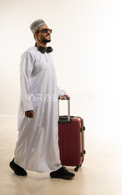 جولة ترفيهية في إجازة الصيف, مفهوم المتعة والحماس، تعابير الفرح والسرور، بورتريه لشاب عربي خليجي عماني مبتسم يرتدي الدشداشة والكمه والنظارة الشمسية وسماعات الرأس يمسك بيده حقيبة السفر, الاستعداد لقضاء رحلة صيفية ممتعة, صورة الجسم بالطول الكامل، خلفية بيضاء