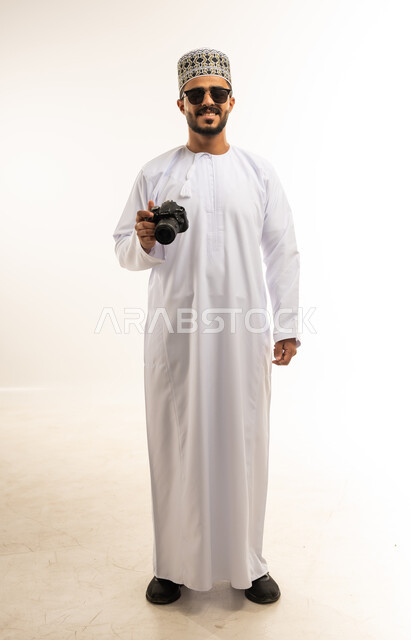 Using high-resolution digital cameras, mastering and professionalizing the photography profession, portrait of a smiling young Arab Gulf Omani man wearing a dishdasha, a sleeve and sunglasses, looking at the camera with gestures of self-confidence, taking souvenir photos using a photographic camera, full-length body image, white background