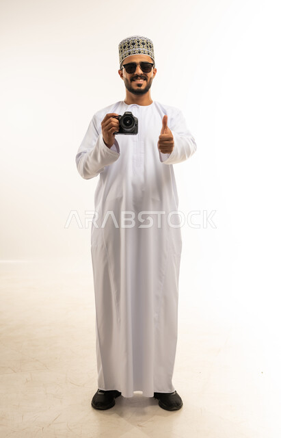 التقاط صور تذكارية عبر كاميرا فوتوغرافية، إتقان واحتراف مهنة التصوير، استخدام كاميرات رقمية عالية الدقة، بورتريه لشاب عربي خليجي عماني مبتسم يرتدي الدشداشة والكمه والنظارة الشمسية يرفع اصبع الابهام بعلامة الاعجاب والرضا، صورة الجسم بالطول الكامل، خلفية بيضاء