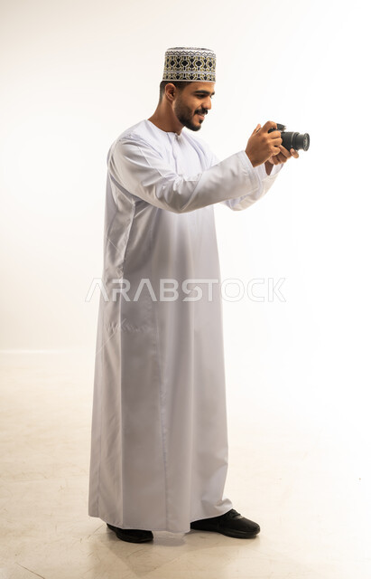 إتقان واحتراف مهنة التصوير، بورتريه من الجانب لشاب عربي خليجي عماني مبتسم يرتدي الدشداشة والكمه يقوم بالتقاط صور تذكارية عبر كاميرا فوتوغرافية، تعابير الفرح والسرور، استخدام كاميرات رقمية عالية الدقة، صورة الجسم بالطول الكامل، خلفية بيضاء