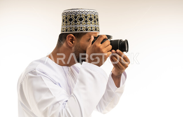 استخدام كاميرات رقمية عالية الدقة، إتقان واحتراف مهنة التصوير، بورتريه مقرب من الجانب لشاب عربي خليجي عماني مبتسم يرتدي الدشداشة والكمه يقوم بالتقاط صور تذكارية عبر كاميرا فوتوغرافية، تعابير الفرح والسرور والسعادة، خلفية بيضاء