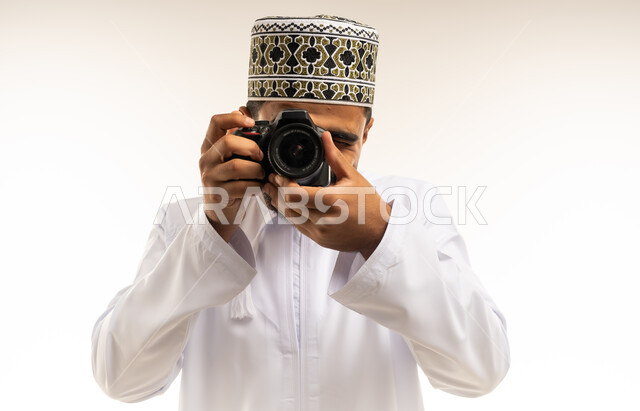 استخدام كاميرات رقمية عالية الدقة، إتقان واحتراف مهنة التصوير، بورتريه مقرب لشاب عربي خليجي عماني مبتسم يرتدي الدشداشة والكمه يقوم بالتقاط صور تذكارية عبر كاميرا فوتوغرافية، تعابير الفرح والسرور والسعادة، خلفية بيضاء