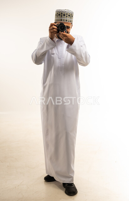 Mastery and professionalism in the photography profession, portrait of a smiling young Arab Gulf Omani man wearing a dishdasha and a kummah taking souvenir photos with a photographic camera, expressions of joy and happiness, using high-resolution digital cameras, full-length body photo, white background