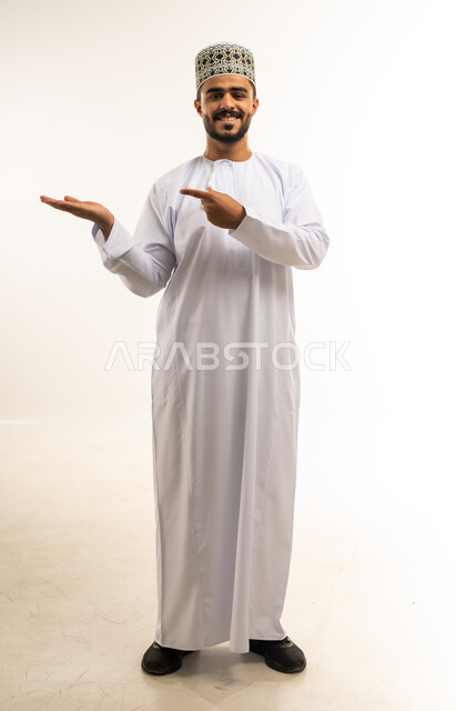 محاولة رفع عنصر ما، مفهوم الاناقة و الاهتمام بالمظهر الخارجي, بورتريه لشاب عربي خليجي عماني مبتسم يرتدي الدشداشة والكمه يشير بإصبع السبابة الى كفه يحمل عناصرهولوجرامية ثلاثية الأبعاد ينظر الى الكاميرا بإيماءات السرور، صورة الجسم بالطول الكامل, خلفية بيضاء