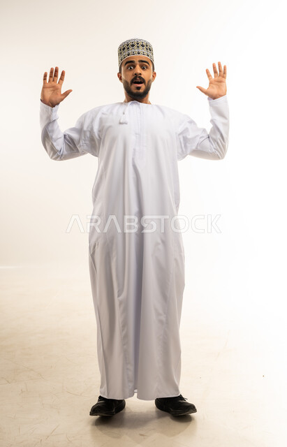 التعجب و التفاجؤ من شيء ما، الشعور بالدهشة والصدمة والذهول, بورتريه لشاب عربي خليجي عماني مبتسم يرتدي الدشداشة والكمه يرفع يديه للاعلى ينظر الى الكاميرا بإيماءات الخوف والذعر, صورة الجسم بالطول الكامل, خلفية بيضاء