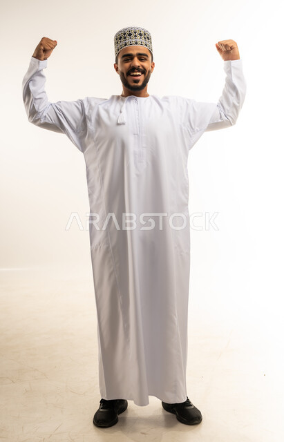 تعابير الفرح والسرور والسعادة، مفهوم البنية الجسدية القوية، اللياقة البدنية والجسم الصحي المتين، بورتريه مقرب لرجل عربي خليجي عماني يرتدي الدشداشة والكمه ينظر الى الكاميرا يقوم بعرض عضلات يديه بإيماءات القوة، صورة الجسم بالطول الكامل، خلفية بيضاء