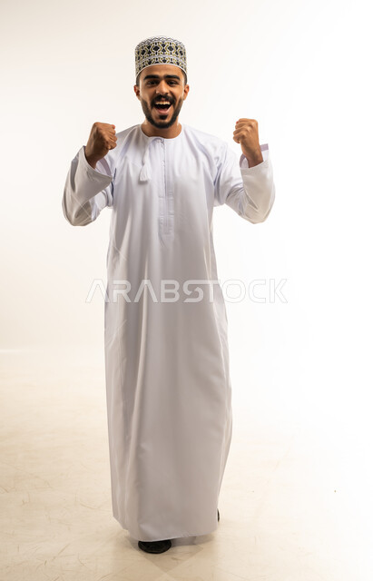 الانتصار والفوز والنجاح، مفهوم الانجاز وتحقيق الاهداف، بورتريه لشاب عربي خليجي عماني مبتسم يرتدي الدشداشة والكمه يرفع قبضتي يديه للاعلى ينظر الى الكاميرا بإيماءات الفرح والسرور, الشعور بالسعادة والقوة، صورة الجسم بالطول الكامل, خلفية بيضاء