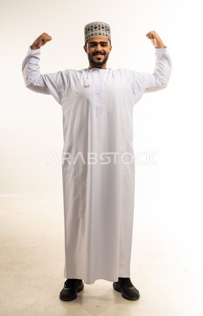 اللياقة البدنية والجسم الصحي المتين، بورتريه مقرب لرجل عربي خليجي عماني يرتدي الدشداشة والكمه يقوم بعرض عضلات يديه ينظر الى الكاميرا بإيماءات القوة، التمتع بالبنية الجسدية القوية، صورة الجسم بالطول الكامل، خلفية بيضاء