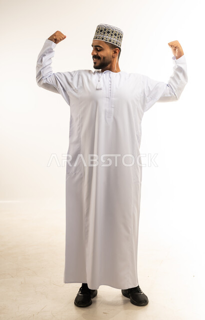 التمتع بالبنية الجسدية القوية، اللياقة البدنية والجسم الصحي المتين، بورتريه مقرب لرجل عربي خليجي عماني يرتدي الدشداشة والكمه يقوم بعرض عضلات يديه بإيماءات القوة، تعابير الغرور والثقة بالنفس، صورة الجسم بالطول الكامل، خلفية بيضاء