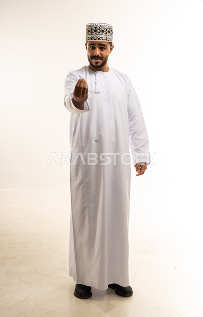 إمهال بعض الوقت للقيام بشئ ما، مفهوم الصبر والتريث، بورتريه لرجل عربي خليجي عماني يرتدي الدشداشة والكمه ينظر الى الكاميرا يشير بيده بإيماءات طلب الإنتظار، مفهوم الصبر والتأن والتريث، صورة الجسم بالطول الكامل، خلفية بيضاء