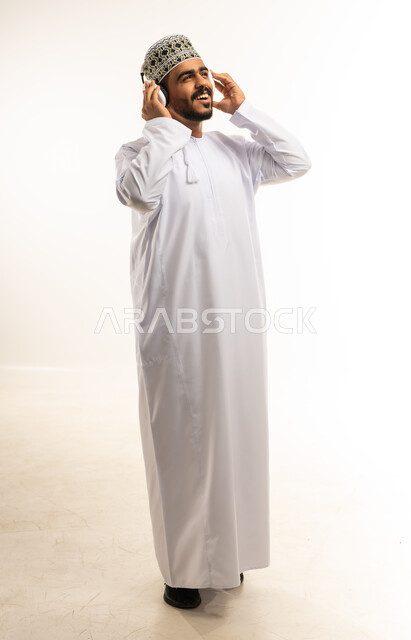 حركات يدين تدل على الرقص والتفاعل مع الموسيقى، بورتريه لشاب عربي خليجي عماني مبتسم يرتدي الدشداشة والكمه يضع سماعات الهيدفون يستمتع بسماع الأغاني المتنوعة، الشعور بالفرح والسعادة، صورة الجسم بالطول الكامل، خلفية بيضاء
