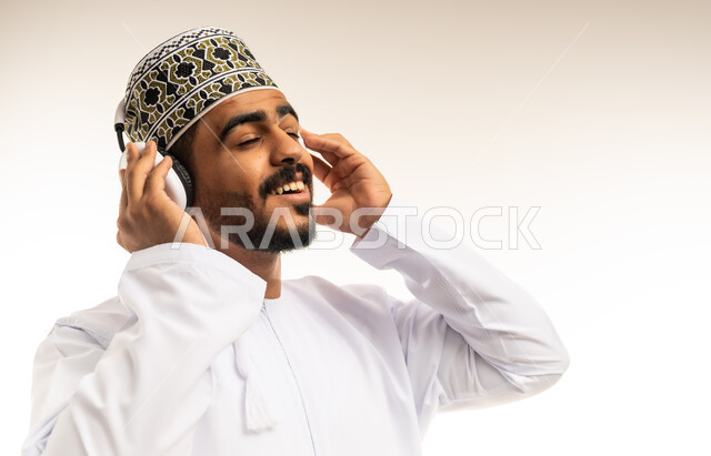 الشعور بالفرح والسعادة، حركات يدين تدل على الرقص والتفاعل مع الموسيقى، بورتريه مقرب لشاب عربي خليجي عماني مبتسم يرتدي الدشداشة والكمه يضع سماعات الهيدفون يستمتع بسماع الأغاني المتنوعة، خلفية بيضاء