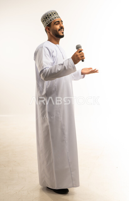 الاندماج والتفاعل مع الموسيقا، تقديم العروض الصوتية، تسجيل مقطع صوتي غنائي، بورتريه لشاب عربي خليجي عماني مبتسم  يرتدي الدشداشة والكمه يمسك بيده المايكروفون يقوم بالغناء بإيماءات الفرح والسرور، صورة الجسم بالطول الكامل، خلفية بيضاء