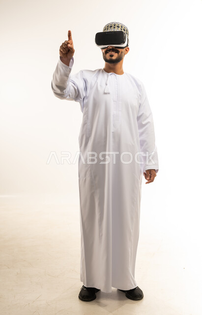 Fusion and simulation of the imaginary world, technical and technological development, clicking with the index finger and interacting with holographic elements, portrait of a young Arab Gulf Omani man wearing a dishdasha and a kimono looking through 3D glasses (VR), full body image, white background