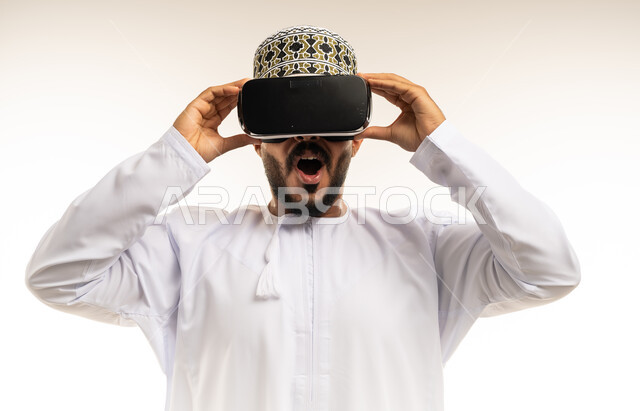 المتعة و التسلية بالمشاهدة عبر نظارة الواقع الافتراضي، التطور التقني والتكنولوجي, بورتريه مقرب لشاب عربي خليجي عماني يرتدي الدشداشة والكمه ينظر من خلال نظارات ثلاثية الأبعاد (الفي ار), الاندماج ومحاكاة العالم الخيالي، تعابير الاندهاش والتفاجؤ، خلفية بيضاء