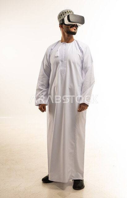Technical and technological development, fun and entertainment by watching through virtual reality glasses, portrait of a young Arab Gulf Omani man wearing a dishdasha and a kimono looking through 3D glasses (VR), integration and simulation of the imaginary world, full body image, white background