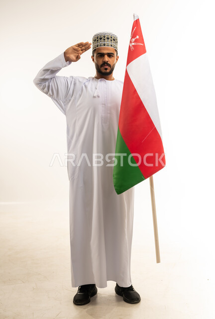مفهوم حب الوطن والإنتماء إليه، الاحتفال بذكرى اليوم الوطني العماني 18 نوفمبر، المناسبات والأعياد الوطنية، بورتريه لشاب عربي خليجي عماني يرتدي الدشداشة والكمه يمسك علم سلطنة عمان ينظر الى الكاميرا يرفع يده بإيماءات التحية والاحترام، صورة الجسم بالكامل، خلفية بيضاء