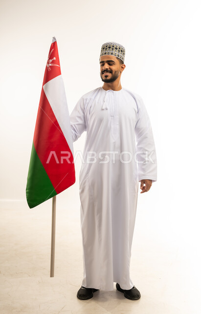 الاحتفال بذكرى اليوم الوطني العماني 18 نوفمبر، المناسبات والأعياد الوطنية، بورتريه لشاب عربي خليجي عماني يرتدي الدشداشة والكمه يقف امام علم سلطنة عمان ينظر اليه بإيماءات الفخر، مفهوم حب الوطن والإنتماء إليه، صورة الجسم بالطول الكامل، خلفية بيضاء