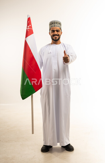 المناسبات والأعياد الوطنية، الاحتفال بذكرى اليوم الوطني العماني 18 نوفمبر، بورتريه لشاب عربي خليجي عماني يرتدي الدشداشة والكمه يقف امام علم سلطنة عمان يرفع اصبع الابهام للاعلى بإيماءات الاعجاب، مفهوم حب الوطن والإنتماء إليه، صورة الجسم بالطول الكامل، خلفية بيضاء