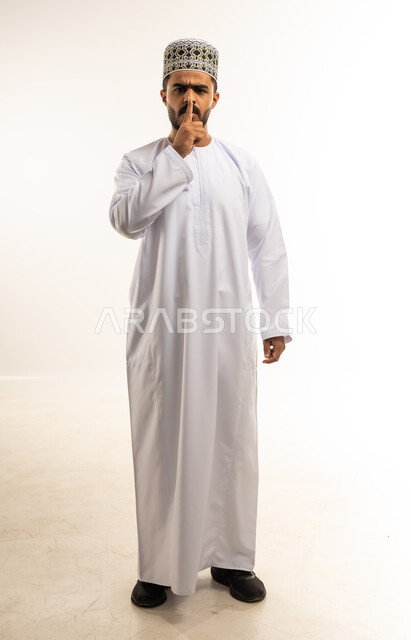 الأمر بعدم الازعاج والسكوت والصمت، بورتريه لشاب عربي خليجي عماني مبتسم يرتدي الدشداشة والكمه يضع اصبع السبابة على فمه ينظر الى الكاميرا بإيماءات التوقف عن الكلام وطلب خفض الصوت، تعابير الجدية والحزم والغضب، صورة الجسم بالطول الكامل، خلفية بيضاء