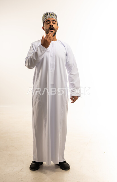 الإحساس بالإرهاق والتعب، بورتريه لشاب عربي خليجي عماني مبتسم يرتدي الدشداشة والكمه يضع يديه على فمه بإيماءات النعاس والحاجة الى النوم والراحة، تعابير التثاؤب والخمول والرغبة في النوم، صورة الجسم بالطول الكامل، خلفية بيضاء