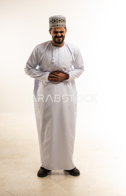 الإحساس بالمغص و التعب والإرهاق, الحاجة للدواء ووضع خطة علاجية, عسر الهضم والشعور بحرقة في المعدة، بورتريه لشاب عربي خليجي عماني مبتسم يرتدي الدشداشة والكمه يضع يديه على بطنه بإيماءات الألم، صورة الجسم بالطول الكامل، خلفية بيضاء