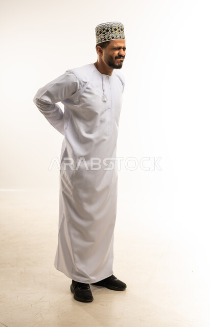 آلام الظهر والعمود الفقري والتهاب المفاصل، بورتريه لشاب عربي خليجي عماني مبتسم يرتدي الدشداشة والكمه يقف بصعوبة ويمسك ظهره بيديه بإيماءات المعاناة، الإحساس بالألم والإصابة بمرض الديسك، صورة الجسم بالطول الكامل، خلفية بيضاء