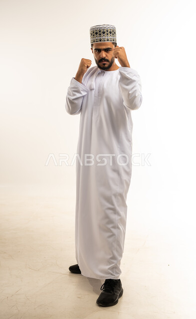 زيادة قدرة العضلات على التحمل، إتقان فنون الملاكمة، بورتريه لشاب عربي خليجي عماني مبتسم يرتدي الدشداشة والكمه يرفع قبضات يديه بإيماءات القوة، ممارسة رياضة الدفاع عن النفس، صورة الجسم بالطول الكامل، خلفية بيضاء