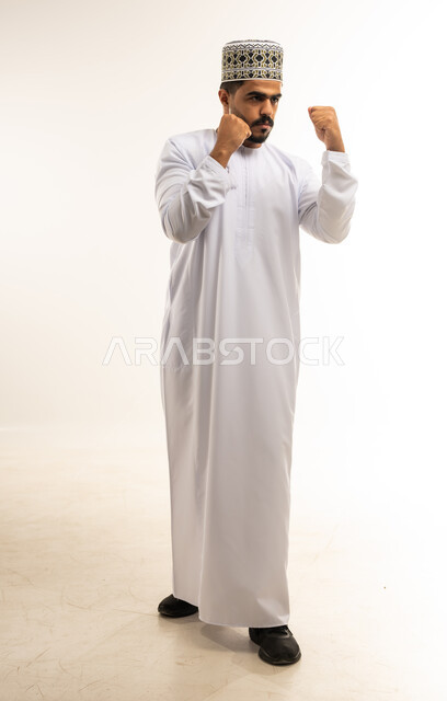 إتقان فنون الملاكمة، زيادة قدرة العضلات على التحمل، بورتريه لشاب عربي خليجي عماني مبتسم يرتدي الدشداشة والكمه يرفع قبضات يديه بإيماءات القوة، ممارسة رياضة الدفاع عن النفس، صورة الجسم بالطول الكامل، خلفية بيضاء