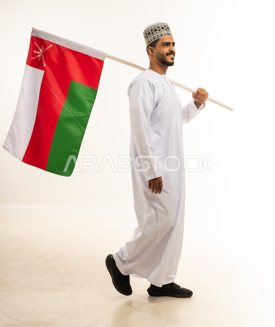 المشاركة بالاعياد والمناسبات الوطنية، الاحتفال بذكرى اليوم الوطني العماني 18 نوفمبر، بورتريه لشاب عربي خليجي عماني يرتدي الدشداشة والكمه يحمل بيده علم سلطنة عمان بإيماءات السرور، مفهوم حب الوطن والإنتماء إليه، الجسم بالطول الكامل، خلفية بيضاء
