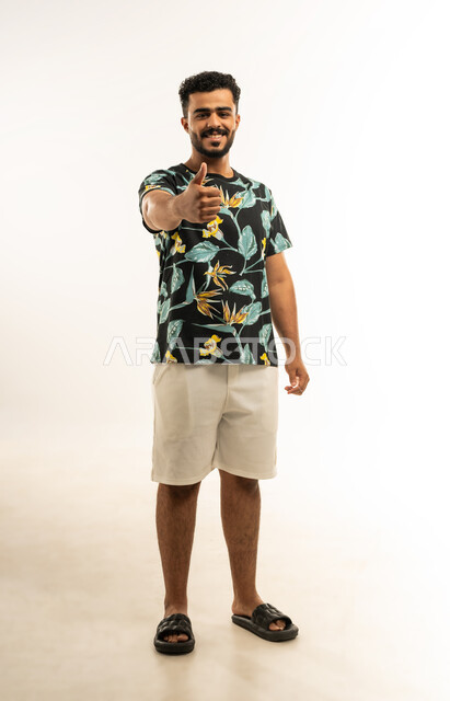 النظر الى الكاميرا بتعابير السعادة والسرور، النجاح وتحقيق الأهداف، علامة الجودة والامتياز, بورتريه لشاب عربي خليجي عماني يرتدي الزي الكاجوال يرفع اصبع الابهام للاعلى بإيماءات الإعجاب والرضا، مفهوم الموافقة والقبول، صورة الجسم بالطول الكامل، خلفية بيضاء