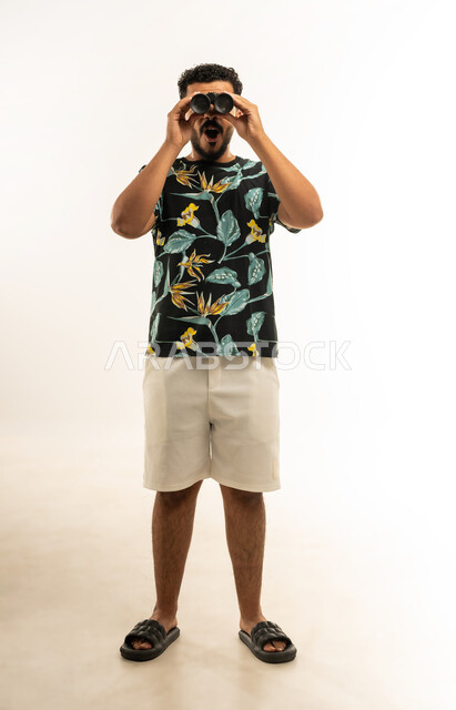 التخطيط المستقبلي للأهداف والطموحات، حب المغامرة والاستكشاف، رؤية الاشياء البعيدة، بورتريه لشاب عربي خليجي عماني مبتسم يرتدي الزي الكاجوال يحمل بيده المنظار ثنائي العينين (التلسكوب) ينظر الى شيء ما بإيماءات الدهشة والتعجب، صورة الجسم بالطول الكامل، خلفية بيضاء