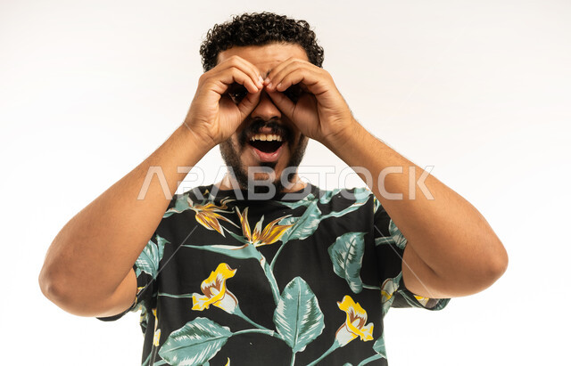 Seeing distant things, love of adventure and exploration, future planning for goals and ambitions, close-up portrait of a smiling young Arab Gulf Omani man wearing casual clothes, putting his hands on his eyes in the shape of binoculars, expressions of joy and happiness, white background