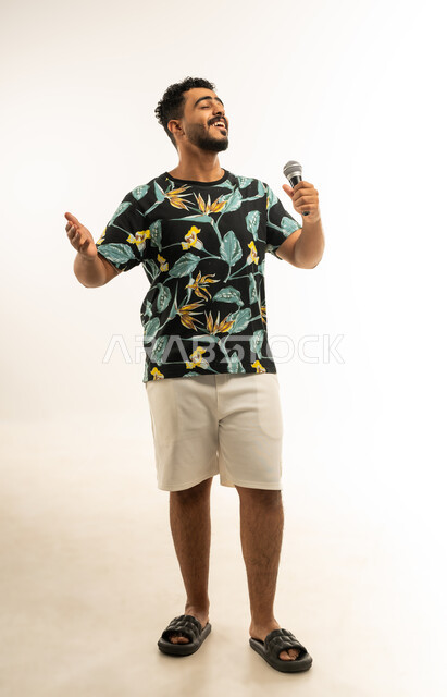 تسجيل مقطع صوتي غنائي، تعابير الاندماج والتفاعل مع الموسيقا، بورتريه مقرب لشاب عربي خليجي عماني مبتسم يرتدي الزي الكاجوال يمسك بيده المايكروفون يقوم بالغناء بإيماءات الفرح والسرور، تقديم العروض الصوتية، صورة الجسم بالطول الكامل، خلفية بيضاء