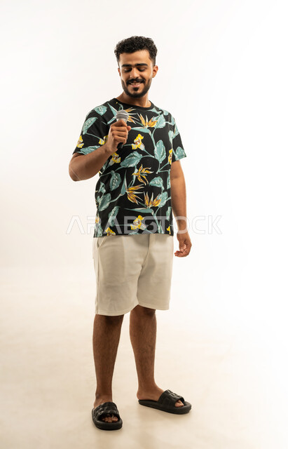 الاندماج والتفاعل مع الموسيقا، تقديم العروض الصوتية، تسجيل مقطع صوتي غنائي، بورتريه لشاب عربي خليجي عماني مبتسم يرتدي الزي الكاجوال يمسك بيده المايكروفون يقوم بالغناء بإيماءات الفرح والسرور، صورة الجسم بالطول الكامل، خلفية بيضاء