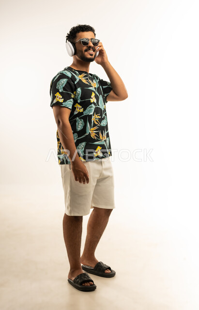 حركات يدين تدل على الرقص والتفاعل مع الموسيقى، بورتريه لشاب عربي خليجي عماني مبتسم يرتدي الزي الكاجوال والنظارة الشمسية يضع سماعات الهيدفون يستمتع بسماع الأغاني المتنوعة، الشعور بالفرح والسعادة، صورة الجسم بالطول الكامل، خلفية بيضاء