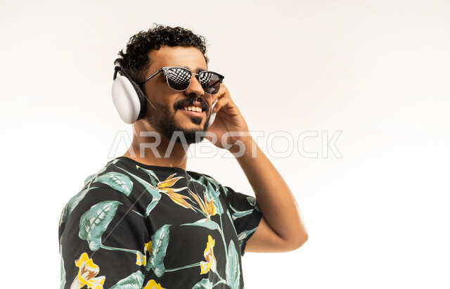 مفهوم الرقص والتفاعل مع الموسيقى، بورتريه مقرب لشاب عربي خليجي عماني مبتسم يرتدي الزي الكاجوال والنظارة الشمسية يضع سماعات الهيدفون يستمتع بسماع الأغاني المتنوعة، الشعور بالفرح والسعادة والسرور، خلفية بيضاء